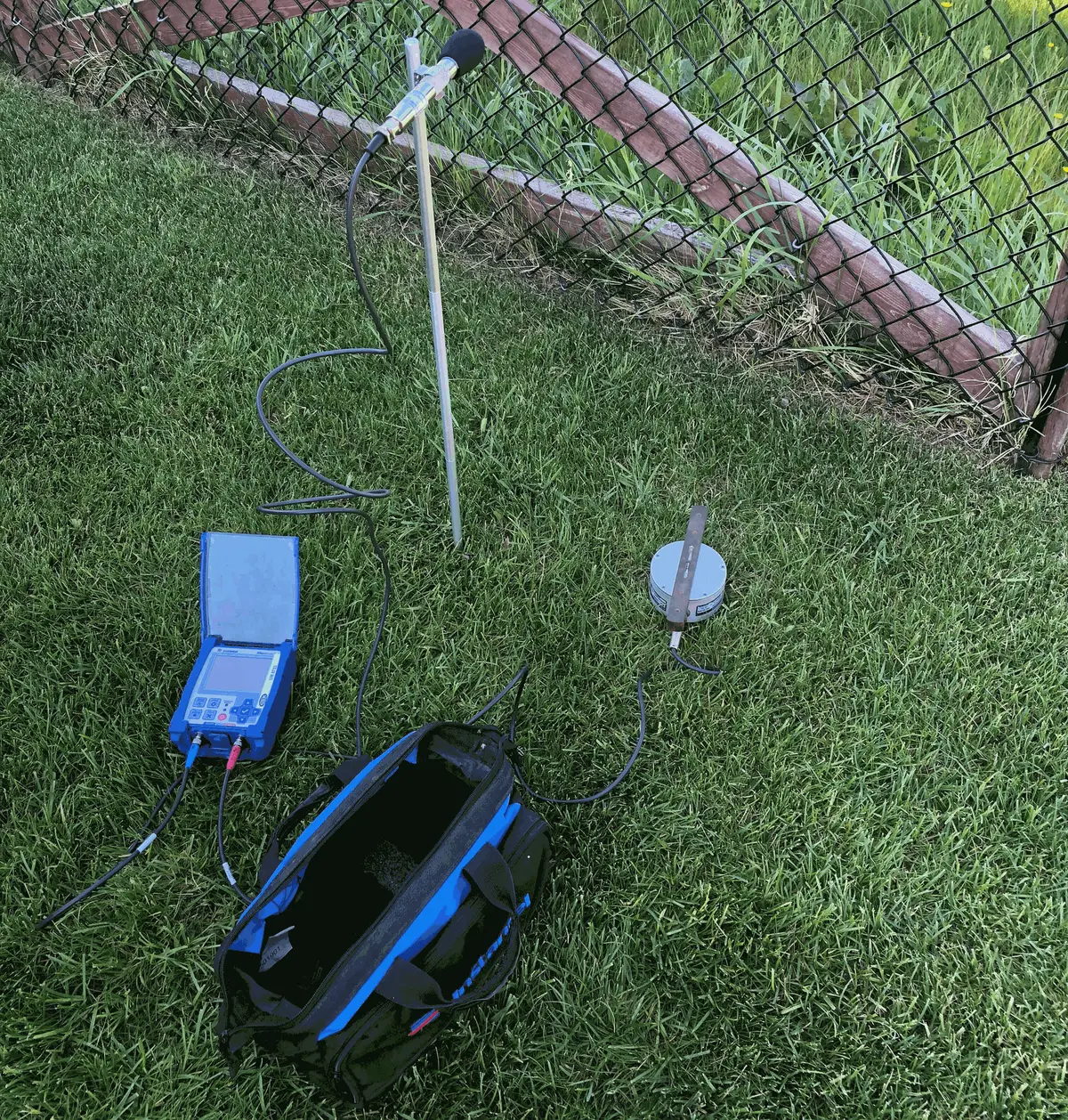 Instantel seismograph on grass beside a fence, with a microphone on a stand and a geophone with a ground spike